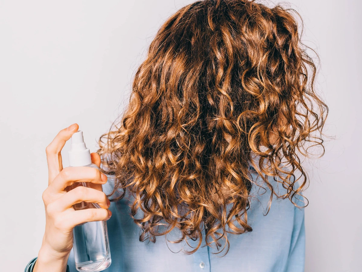 Beach Waves Frisur Junge Frau mit langen lockigen Haaren, die kosmetische Pflegeprodukte aufträgt. Spray mit Meersalz, um Beachy Waves Frisur zu machen.